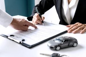Person signing insurance or legal documents, guided by another, with model car nearby.