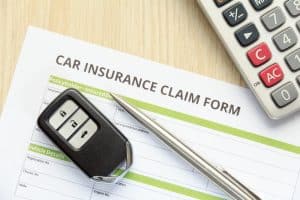 A car insurance claim form, along with a car key and calculator, seen from above, placed on a wooden desk.