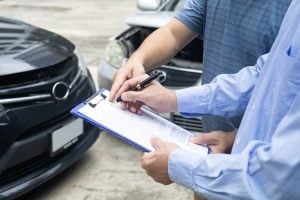 Insurance adjuster and client filling out claim forms by damaged cars after an accident.